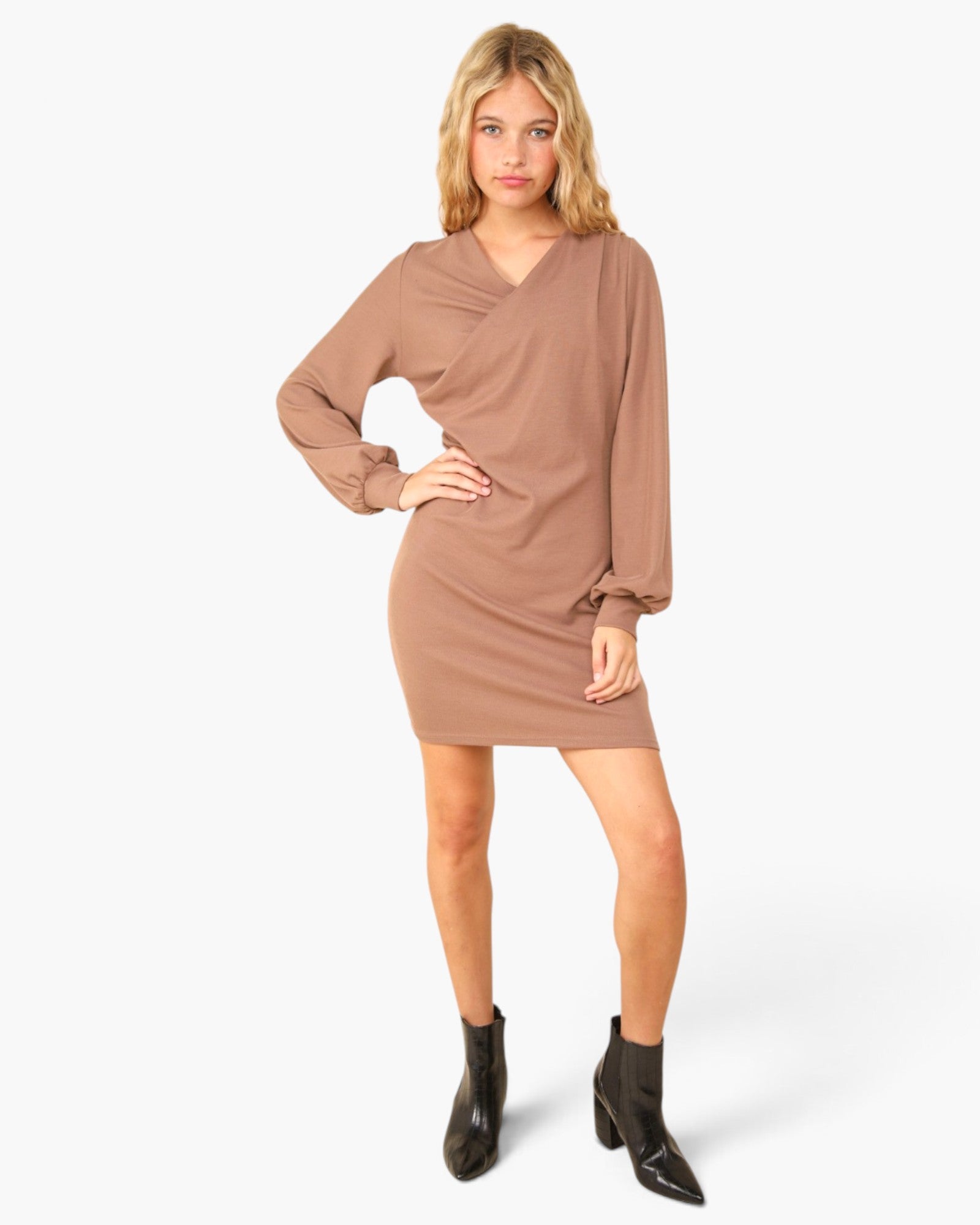 A woman modeling a long-sleeve, wrap-style mini dress in a muted taupe-brown color, paired with black ankle boots, against a plain light gray studio background.