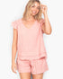 A woman modeling a light pink two-piece lounge set consisting of a short-sleeve V-neck top with ruffle details and matching shorts, set against a plain light gray background.
