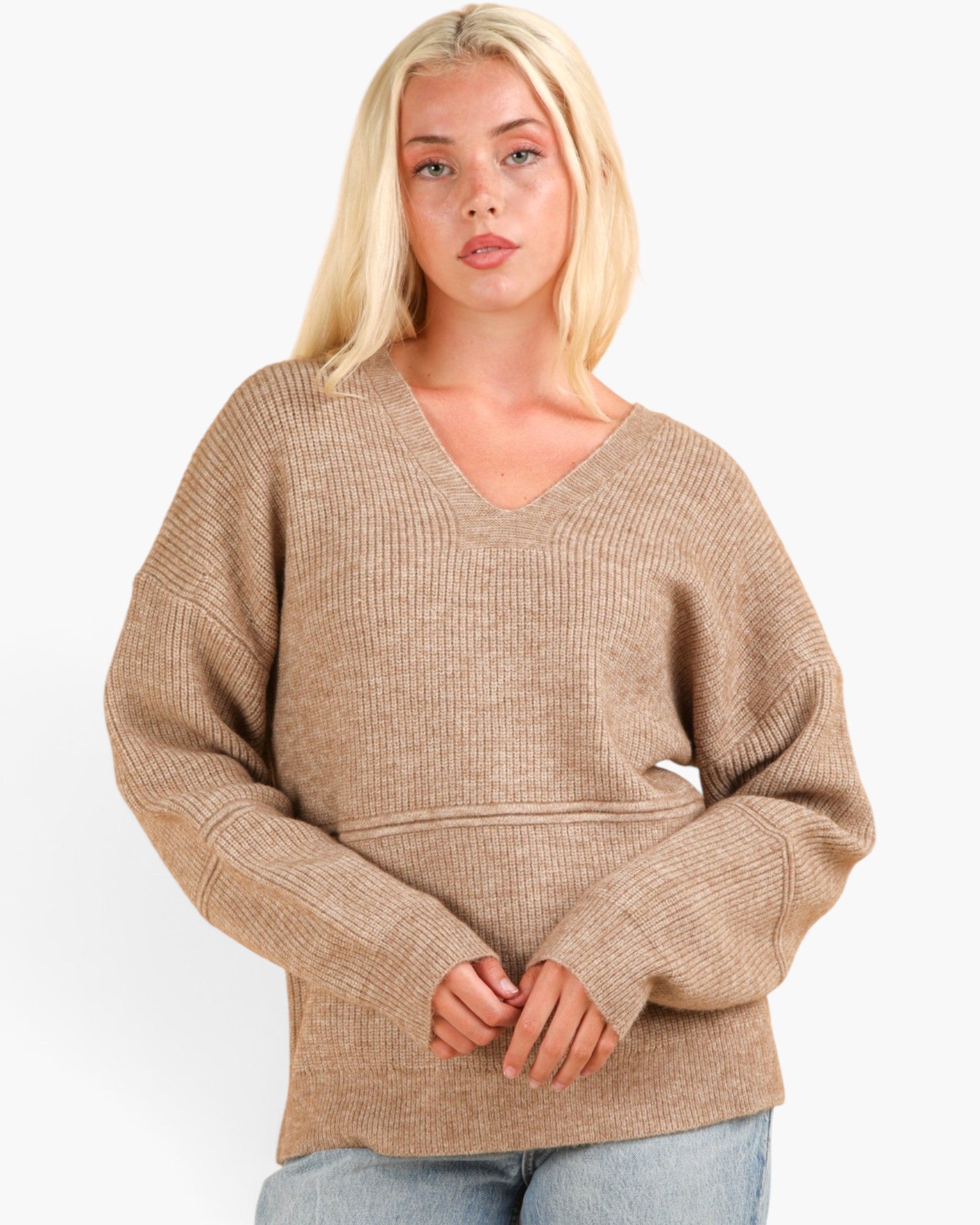 A blonde woman wearing a beige ribbed knit V-neck sweater with oversized sleeves, paired with light blue jeans, against a plain white background.