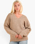 A blonde woman wearing a beige ribbed knit V-neck sweater with oversized sleeves, paired with light blue jeans, against a plain white background.