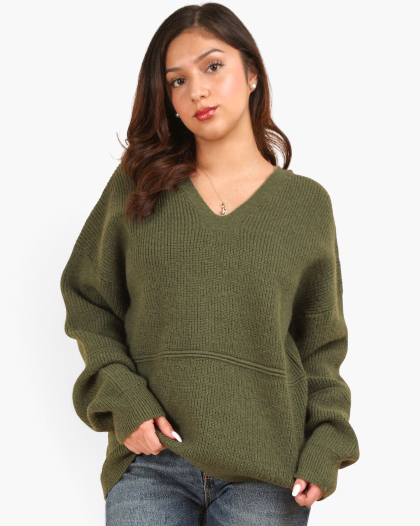A woman wearing an oversized olive green ribbed knit sweater with a V-neck and front pockets, paired with blue jeans, against a plain light gray background.