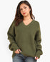 A woman wearing an oversized olive green ribbed knit sweater with a V-neck and front pockets, paired with blue jeans, against a plain light gray background.