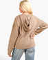 A woman shown from the back wearing a beige ribbed knit hooded sweater and light blue jeans against a plain white studio background.