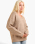 A blonde woman wearing an oversized ribbed knit sweater in a warm beige/tan color, paired with light blue jeans, photographed against a plain light gray studio background.