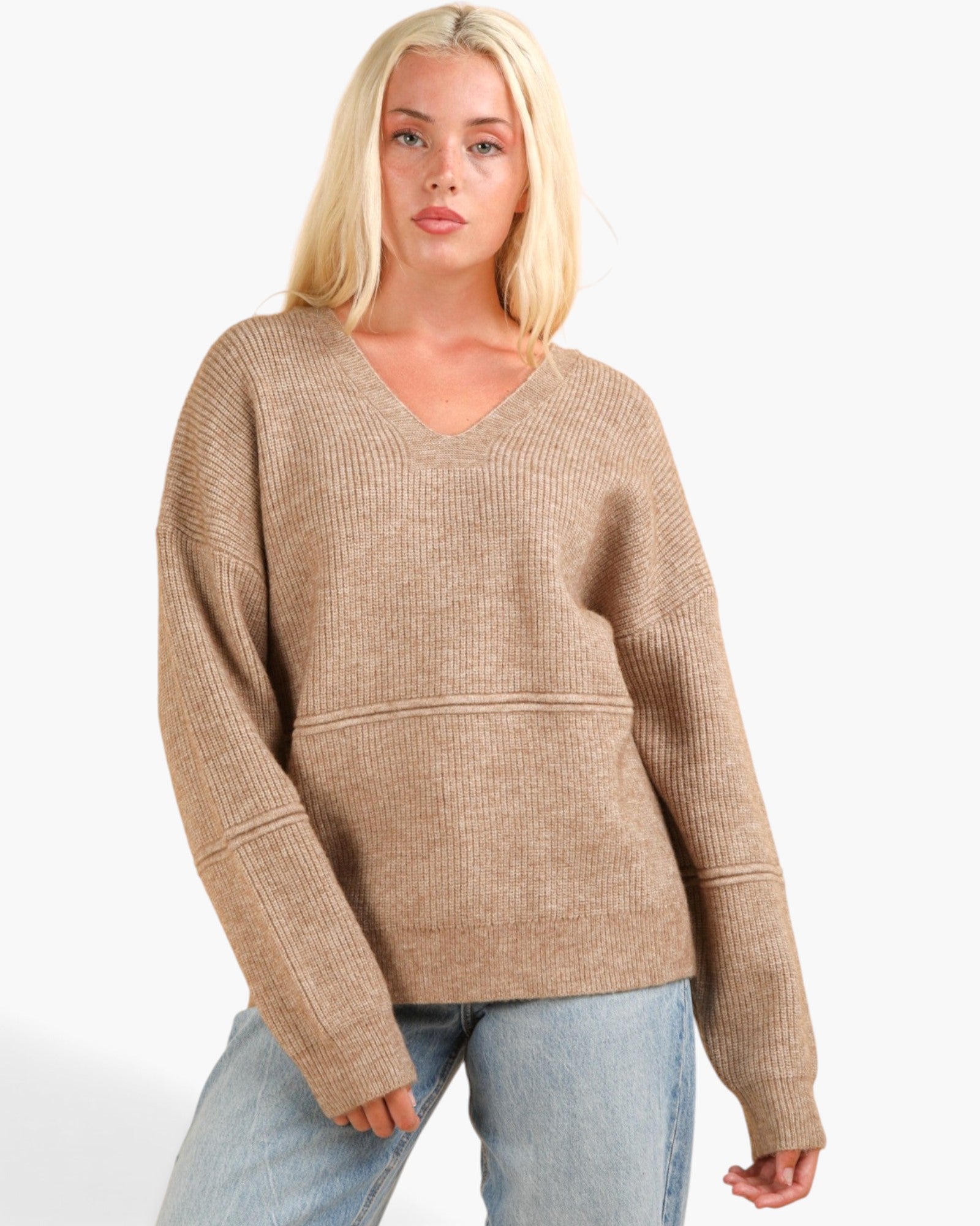 A blonde woman wearing a beige ribbed knit V-neck sweater with long sleeves and light blue jeans, photographed against a plain light gray studio background.