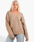 A blonde woman wearing a beige ribbed knit V-neck sweater with long sleeves and light blue jeans, photographed against a plain light gray studio background.