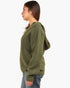 A woman shown in profile wearing an olive green ribbed knit sweater and blue jeans against a plain light gray background.