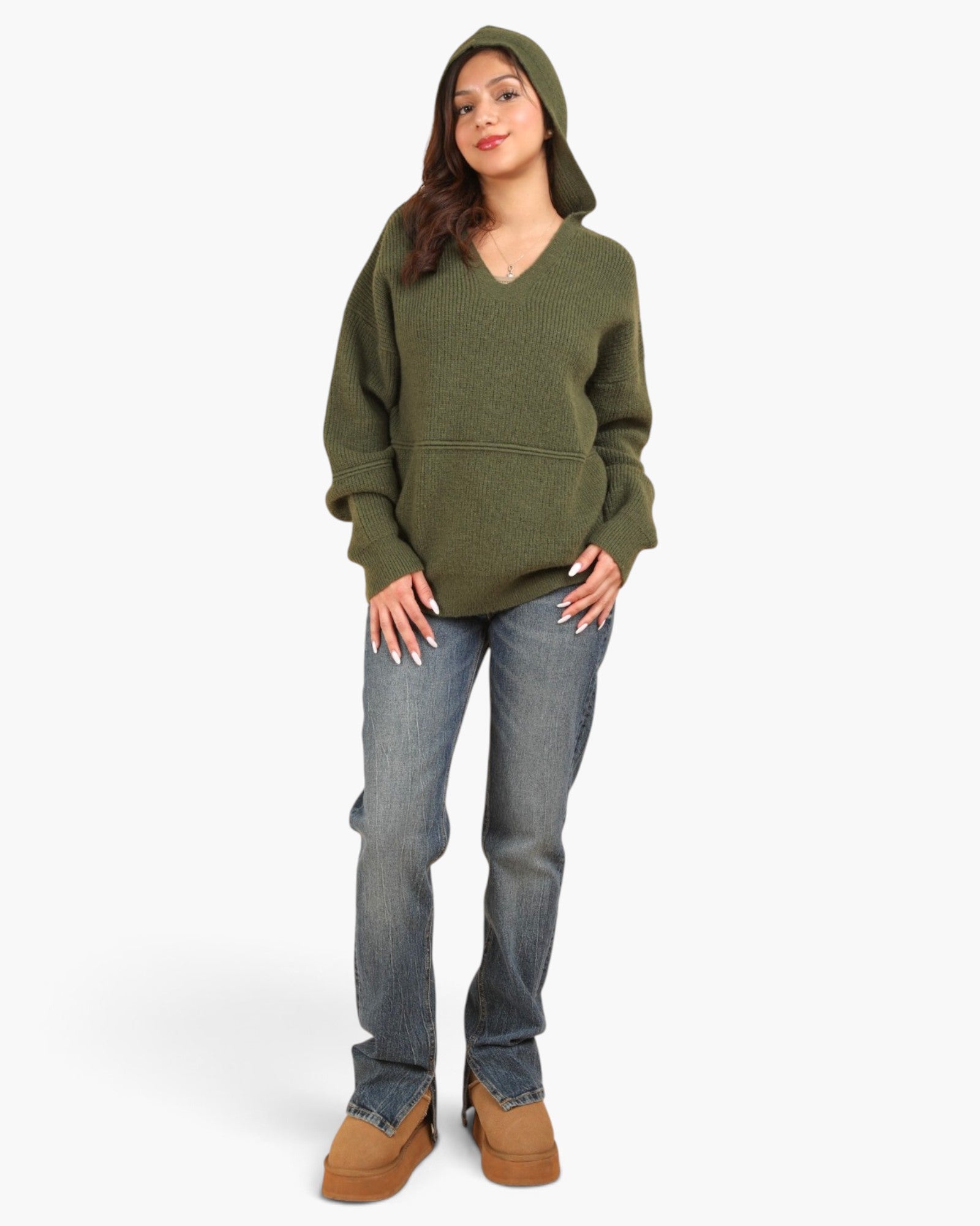 A woman modeling an oversized olive green knit hoodie with a V-neck and front pouch pocket, paired with blue jeans and brown boots, against a plain light gray studio background.