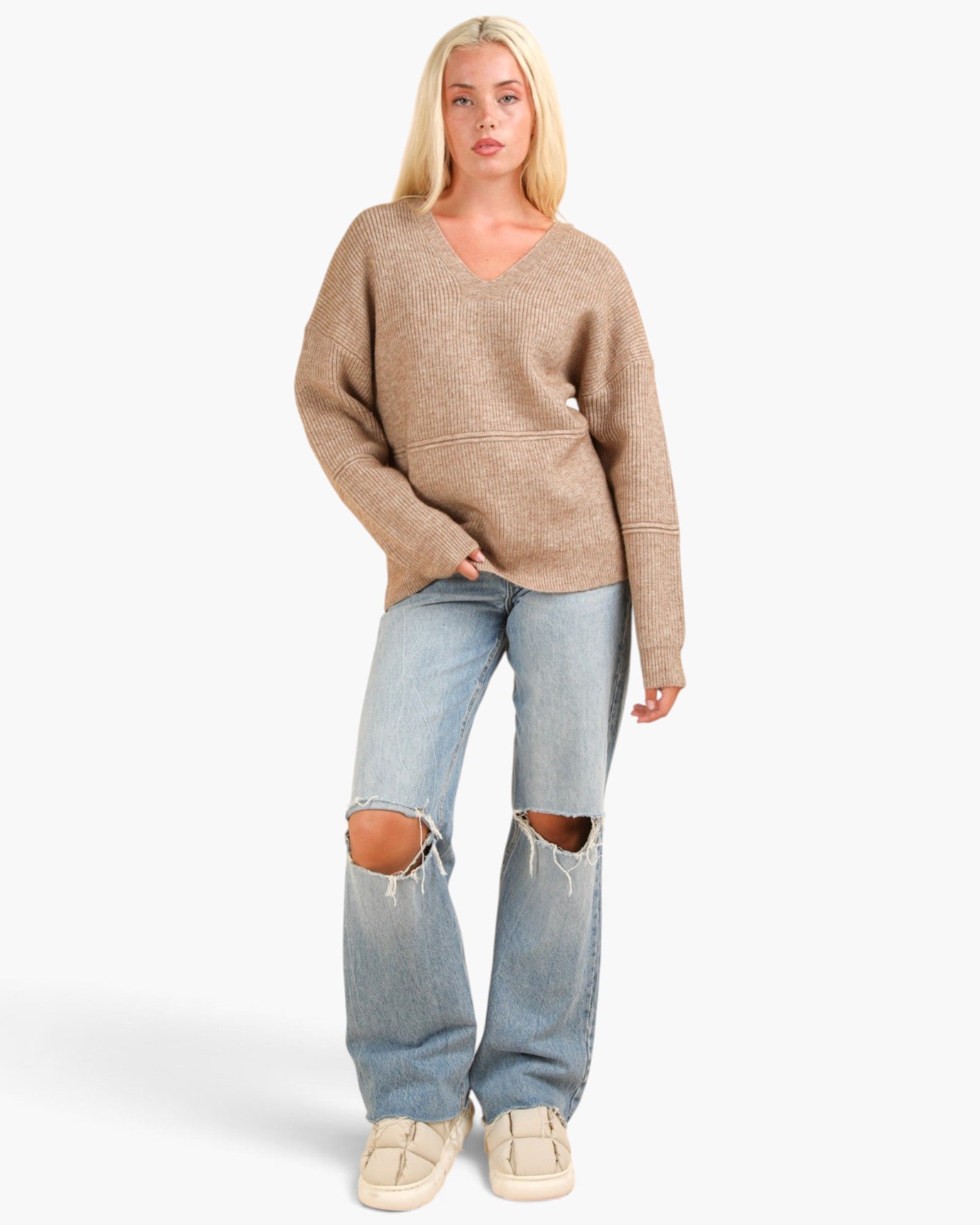 A blonde model wearing a beige knit V-neck sweater, light-wash distressed jeans with ripped knees, and cream sneakers, standing against a light gray studio background.