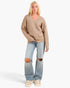 A blonde model wearing a beige knit V-neck sweater, light-wash distressed jeans with ripped knees, and cream sneakers, standing against a light gray studio background.