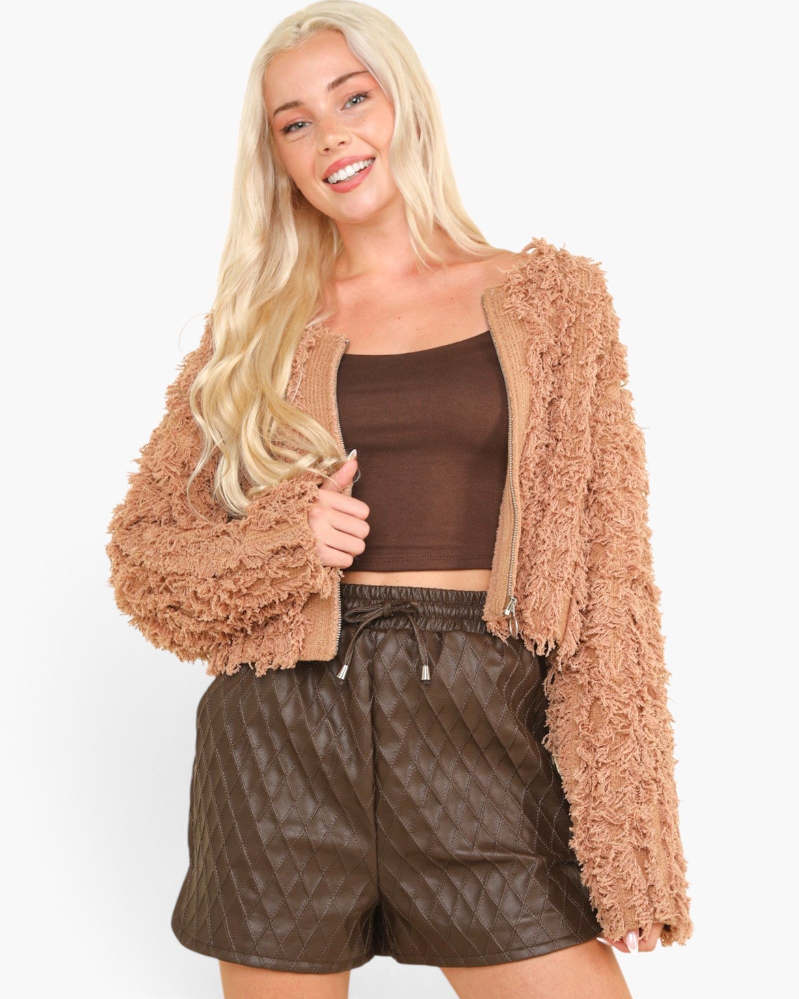 A blonde model wearing a tan fuzzy cropped cardigan over a brown crop top and brown quilted faux-leather shorts, set against a plain white studio background.