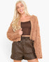 A blonde model wearing a tan fuzzy cropped cardigan over a brown crop top and brown quilted faux-leather shorts, set against a plain white studio background.