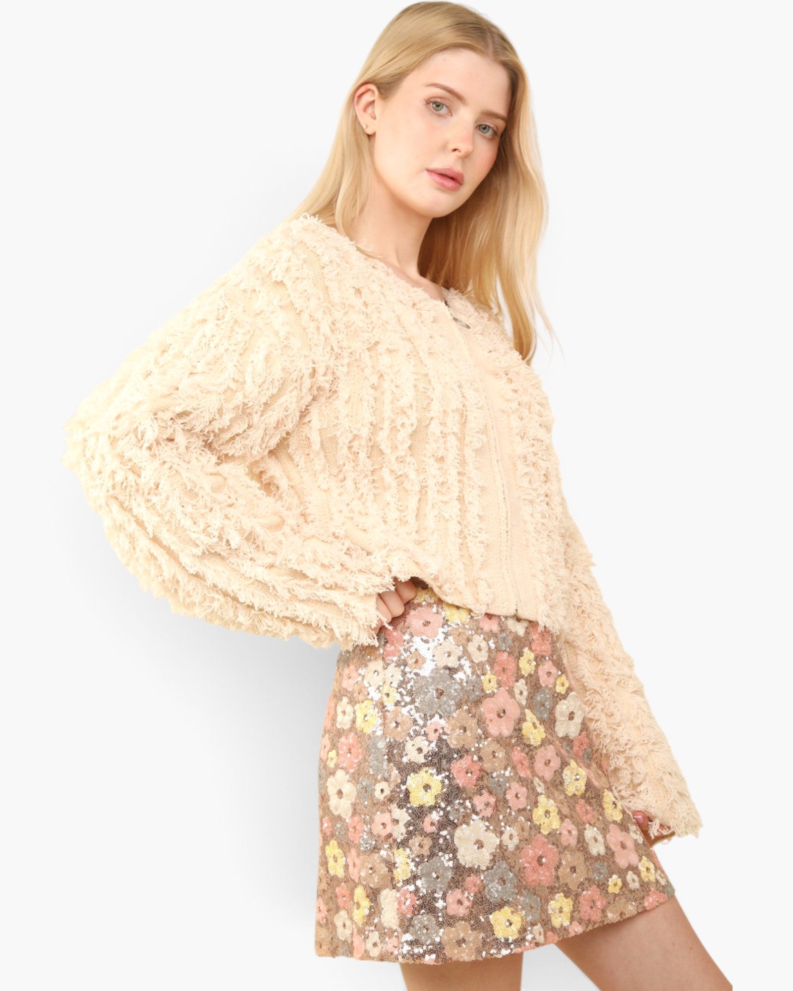 A blonde model wearing a cream-colored, heavily textured ruffled long-sleeve sweater and a sequined mini skirt with a pastel floral pattern, set against a plain light gray background.