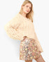 A blonde model wearing a cream-colored, heavily textured ruffled long-sleeve sweater and a sequined mini skirt with a pastel floral pattern, set against a plain light gray background.