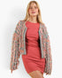 A woman modeling a textured, fringed open-front jacket in a dusty pink, blue, and gray colorway over a solid coral pink dress, set against a plain white studio background.