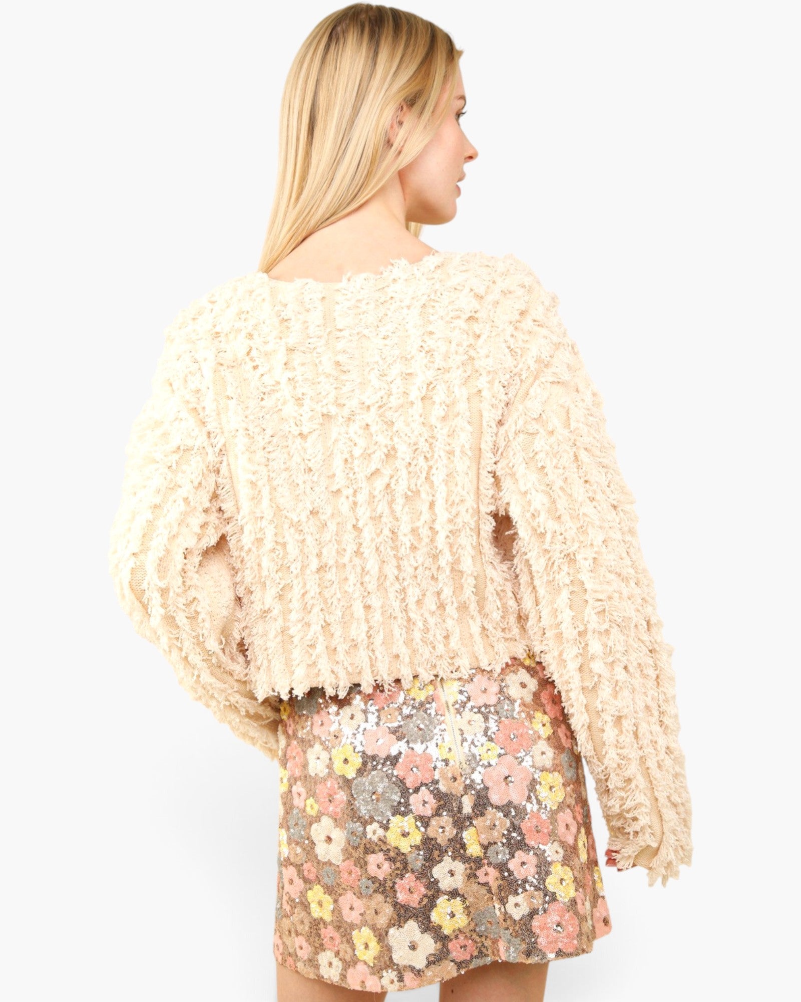 A woman shown from the back wearing a cream-colored, heavily textured ruffled knit sweater and a pink, yellow, and white floral sequined mini skirt, set against a plain light gray studio background.