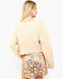A woman shown from the back wearing a cream-colored, heavily textured ruffled knit sweater and a pink, yellow, and white floral sequined mini skirt, set against a plain light gray studio background.