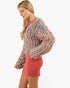 A woman wearing a chunky, fuzzy knit cardigan in a heathered pink, gray, and blue colorway, paired with a coral pink mini dress, photographed against a solid white background.