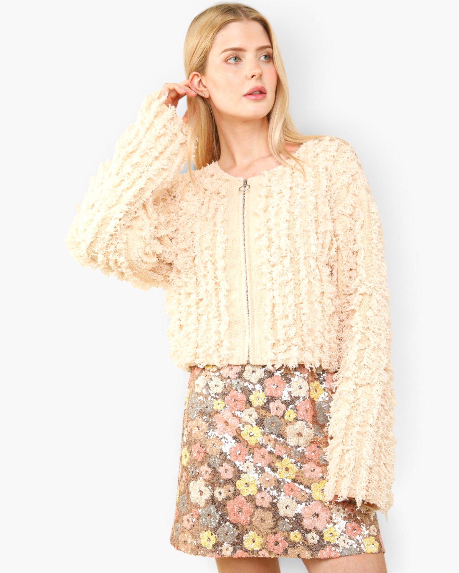 A woman modeling a cream-colored, textured ruffled zip-up jacket paired with a floral sequined mini skirt against a light gray studio background.
