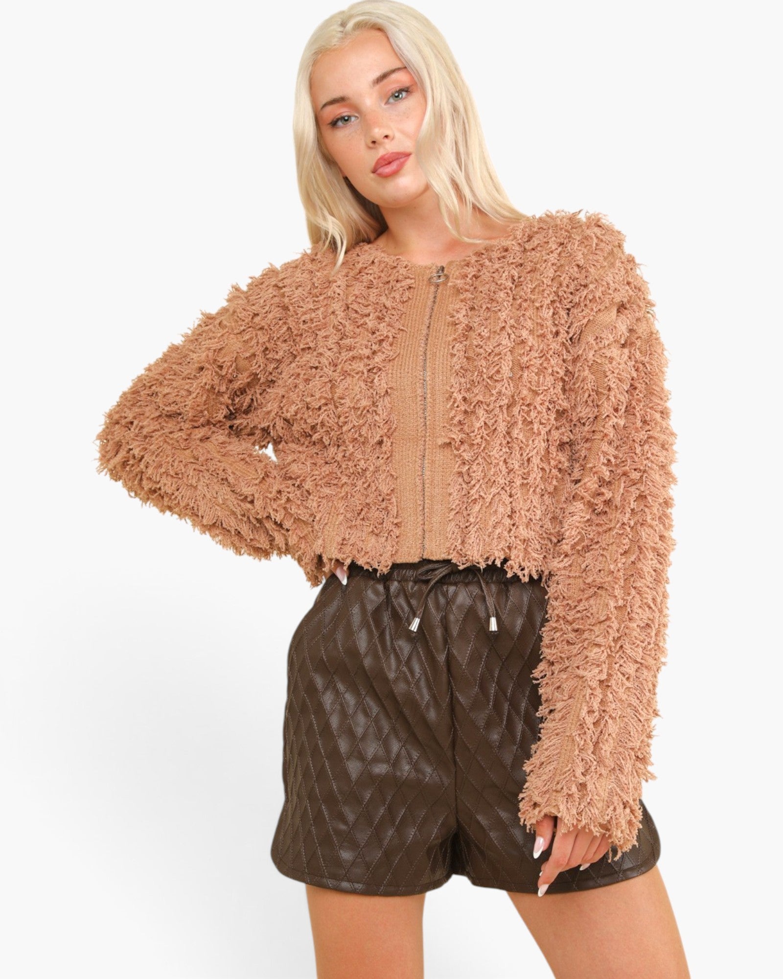 A blonde model wearing a cropped, long-sleeve fuzzy zip-up jacket in a warm tan-brown color, paired with dark brown quilted shorts, against a plain white background.
