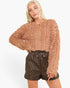 A blonde model wearing a cropped, long-sleeve fuzzy zip-up jacket in a warm tan-brown color, paired with dark brown quilted shorts, against a plain white background.