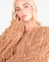 A close-up of a blonde woman wearing a chunky, fringed knit sweater in a warm tan or camel color against a plain light background.