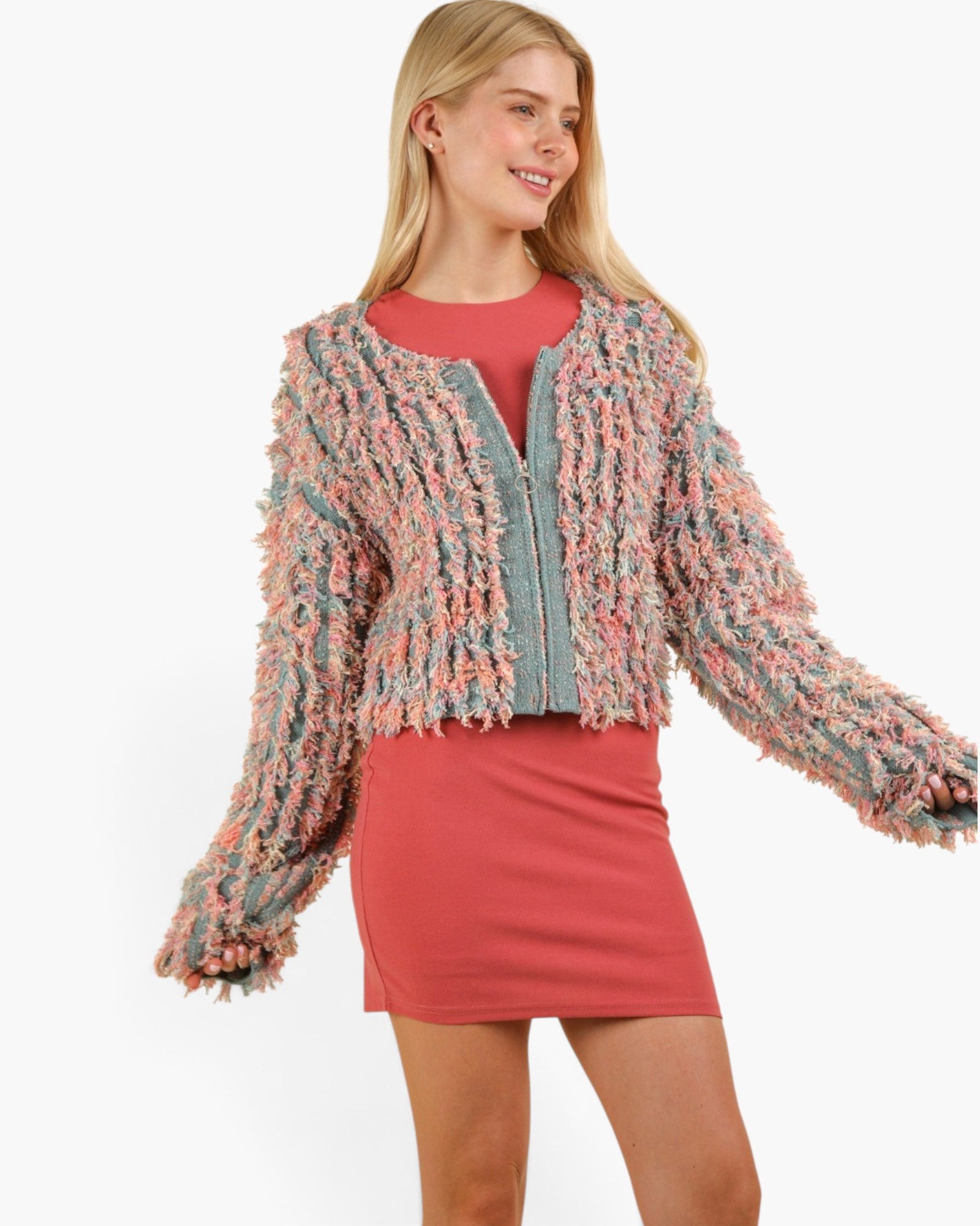 A woman modeling a fuzzy, textured knit cardigan in a pink, peach, and sage green marled pattern, worn over a coral-red top and matching skirt against a plain white background.