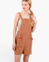 A woman modeling a rust-orange short bib jumpsuit with a subtle grid pattern, worn over a beige tank top against a plain white studio background.