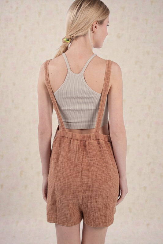 Woman wearing a beige tank top and brown checkered overalls against a light background