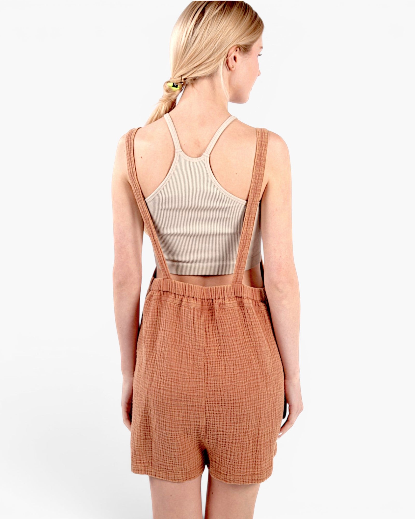 A woman shown from the back wearing a rust-orange textured overall romper over a beige ribbed tank top, against a plain white studio background.