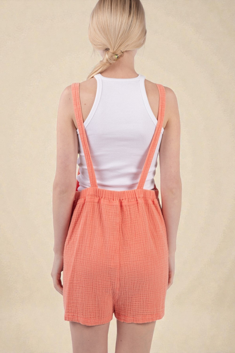 Person wearing a white tank top and coral checkered shorts with suspenders on a beige background