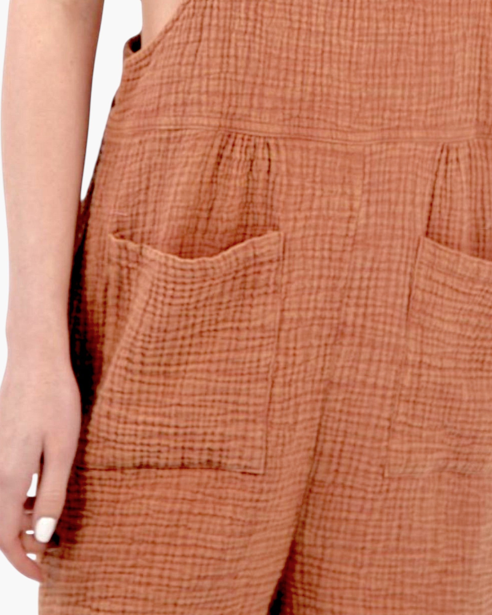 Close-up of a person wearing textured terracotta-colored cotton gauze overalls with front patch pockets and a gingham-like woven pattern.
