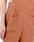 Close-up of a person wearing textured terracotta-colored cotton gauze overalls with front patch pockets and a gingham-like woven pattern.