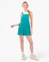 A woman modeling a sleeveless teal corduroy romper with front patch pockets, worn over a white top and paired with white socks and beige sneakers, on a light gray studio background.