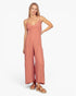A woman modeling a dusty rose wide-leg jumpsuit with thin spaghetti straps and a smocked bodice, paired with brown espadrille sandals, against a plain white studio background.