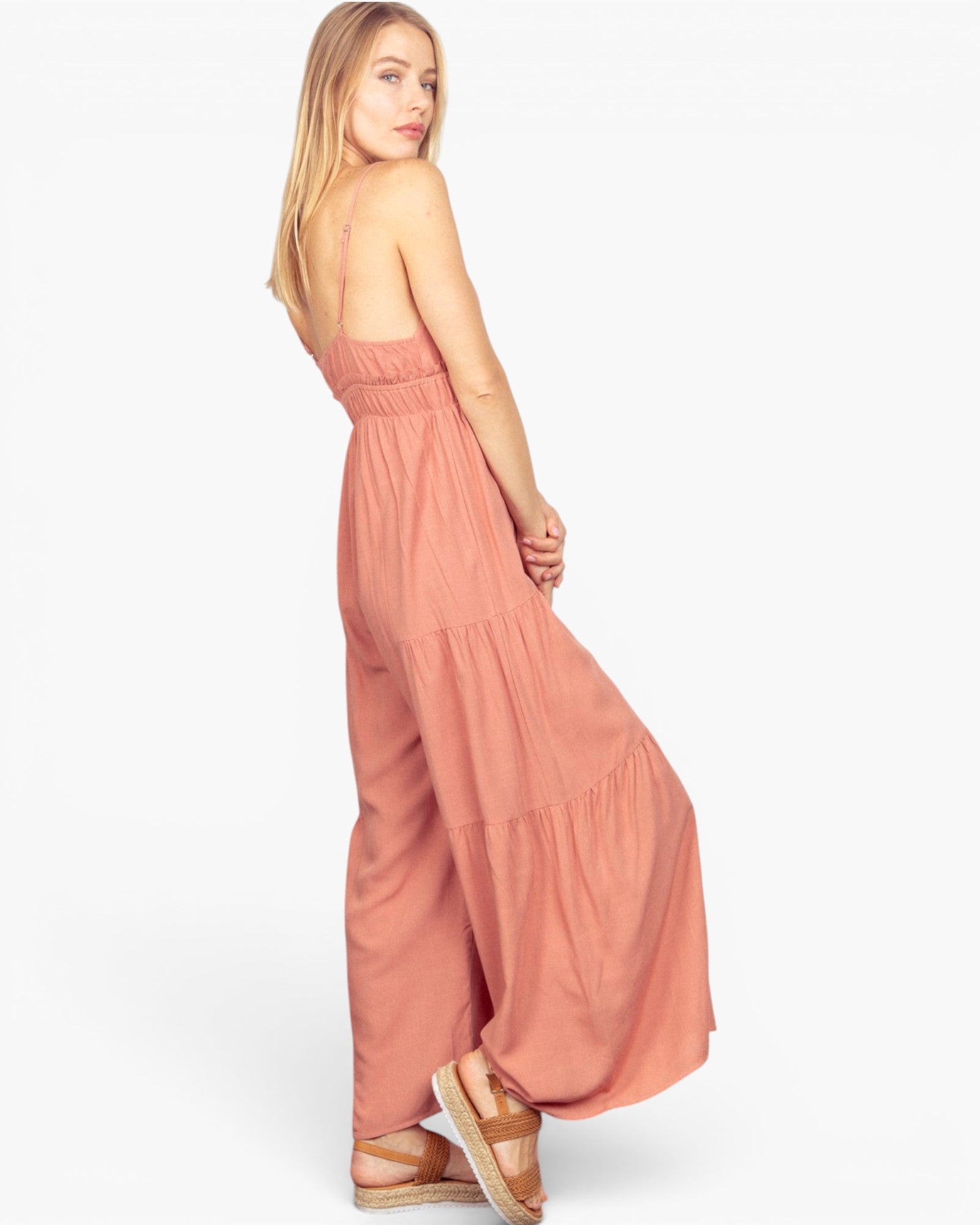 A woman modeling a dusty rose strapless maxi dress with a smocked waistband and tiered, wide-leg pants, paired with brown espadrille sandals against a plain white background.