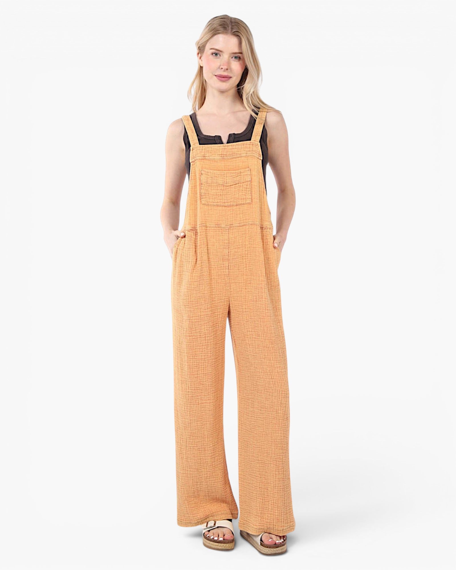 A woman modeling a pair of wide-leg jumpsuit in a textured mustard yellow fabric, styled over a black camisole and brown sandals, against a plain white studio background.