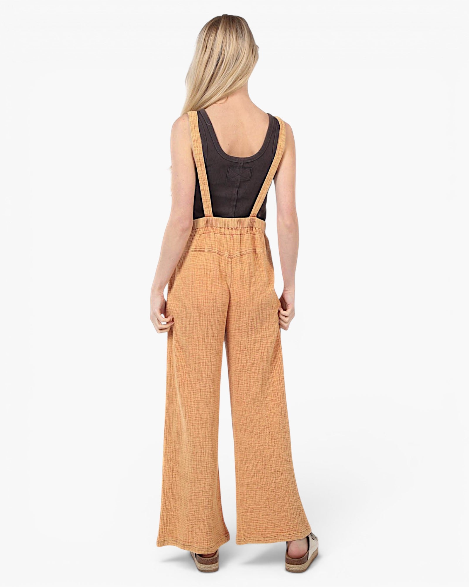 A woman shown from the back wearing a mustard yellow wide-leg jumpsuit with suspenders over a black tank top, set against a plain white studio background.