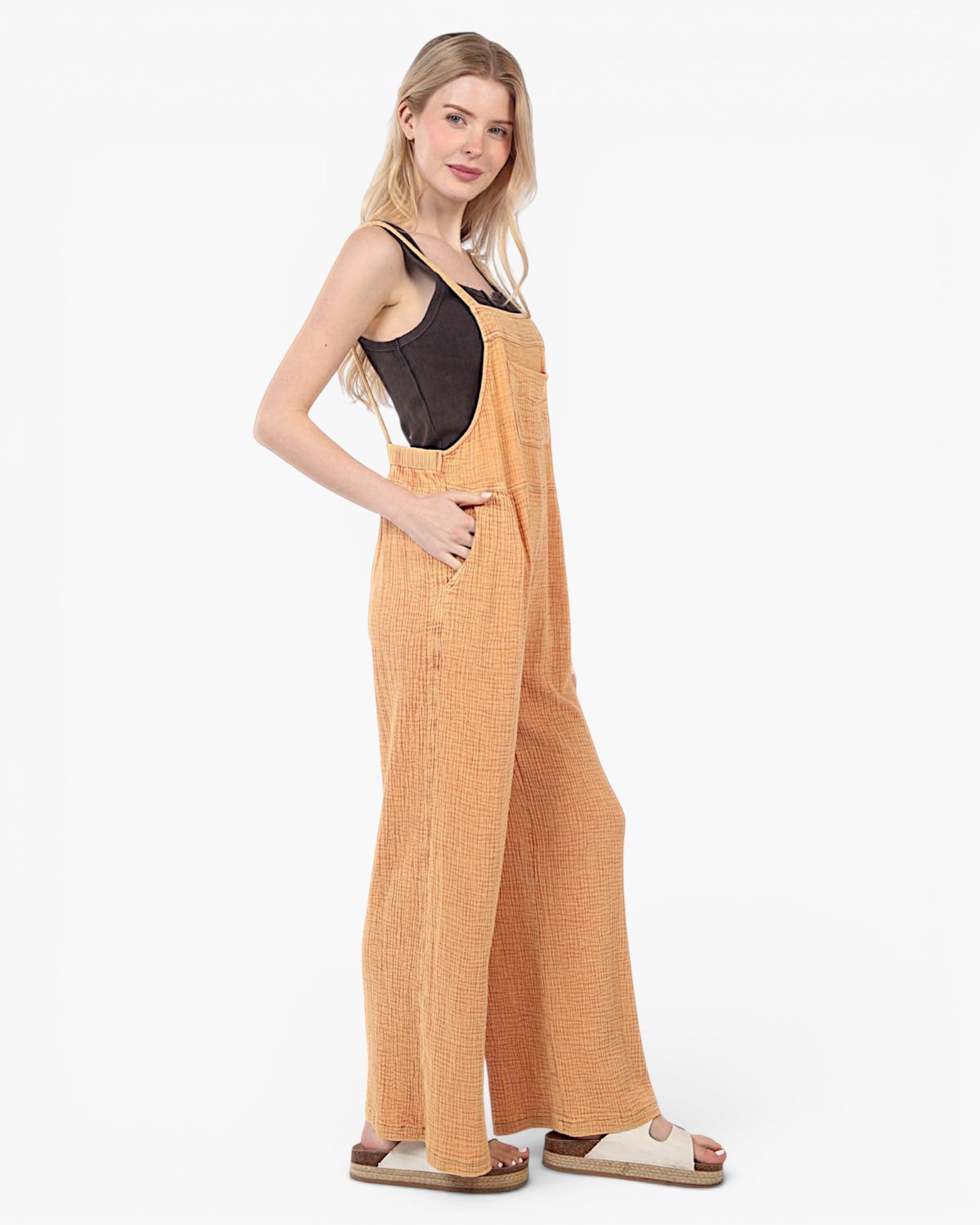 A woman modeling a pair of wide-leg overall jumpsuits in a textured mustard yellow fabric, styled with a black camisole top and beige slide sandals against a plain white studio background.