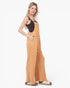 A woman modeling a pair of wide-leg overall jumpsuits in a textured mustard yellow fabric, styled with a black camisole top and beige slide sandals against a plain white studio background.