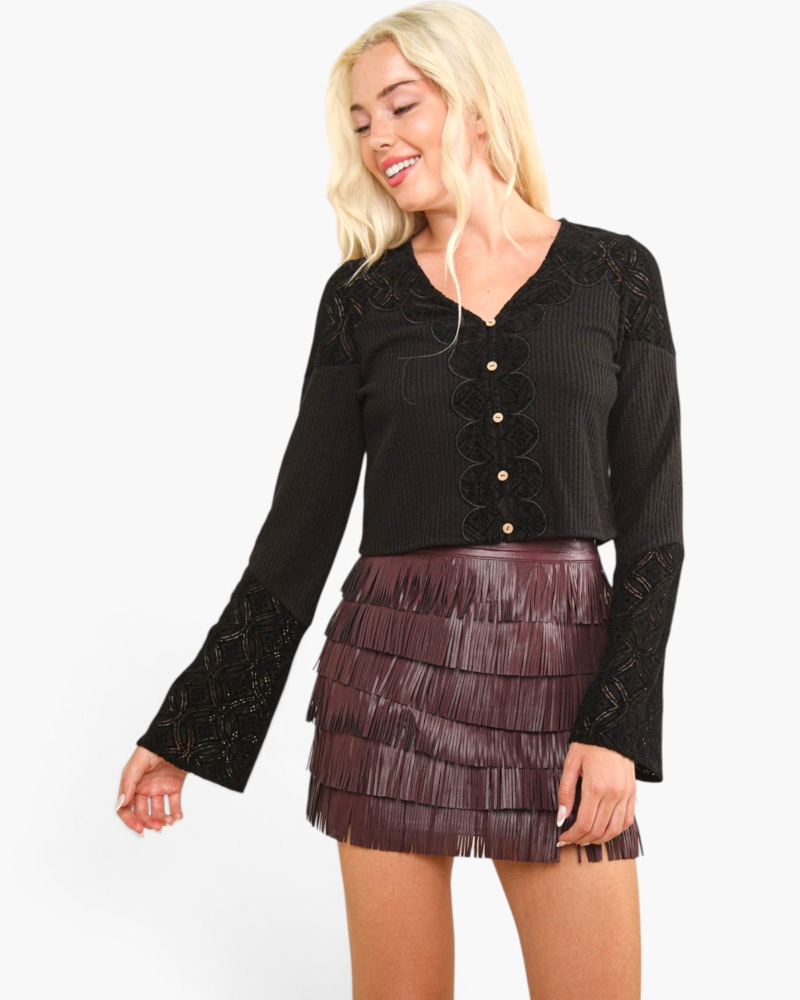A woman modeling a black long-sleeve cropped top with scalloped lace sleeves and a dark brown fringed mini skirt against a plain white background.