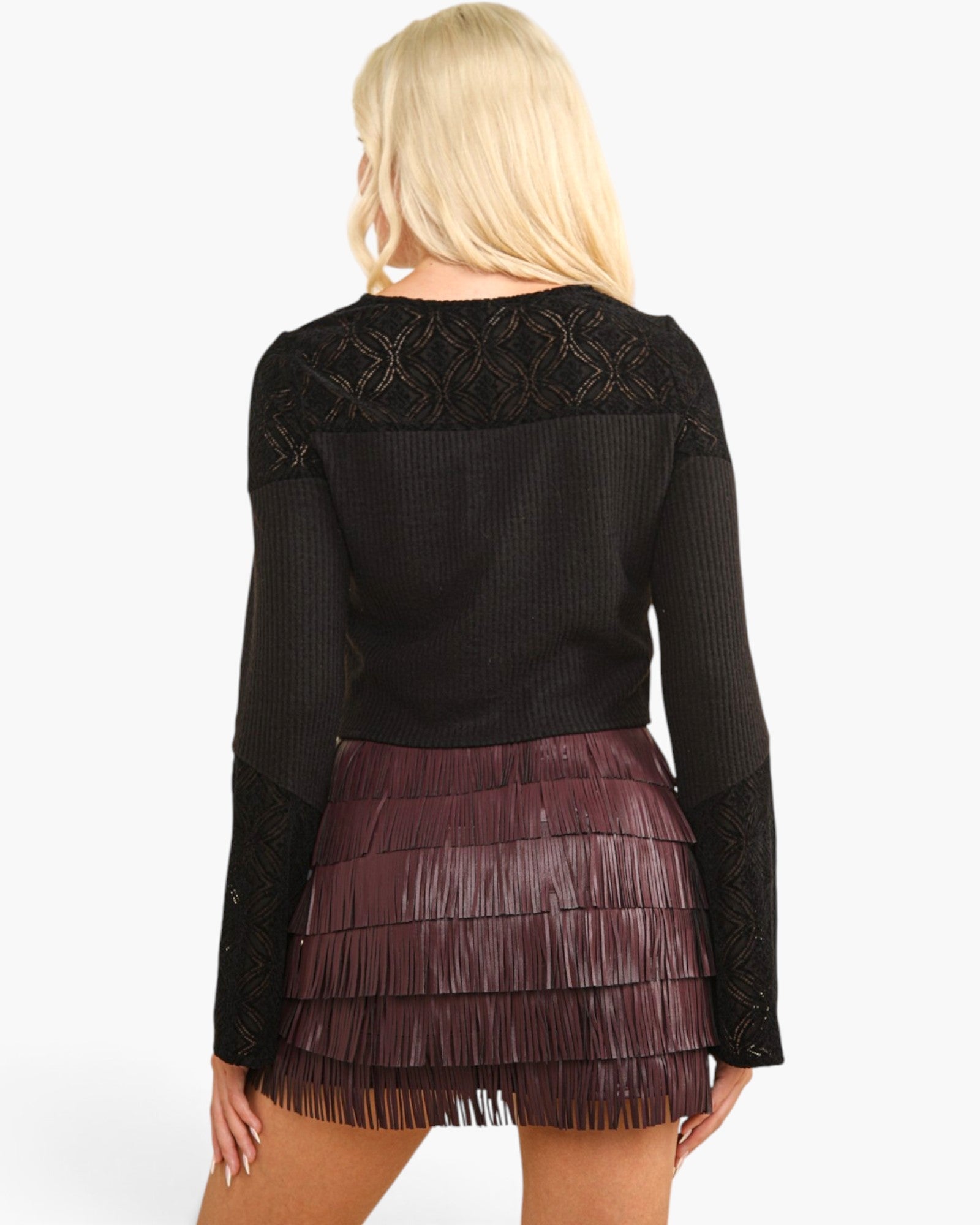 A woman shown from the back wearing a black long-sleeve ribbed knit top with lace shoulder panels and a dark burgundy fringed mini skirt, against a plain white background.