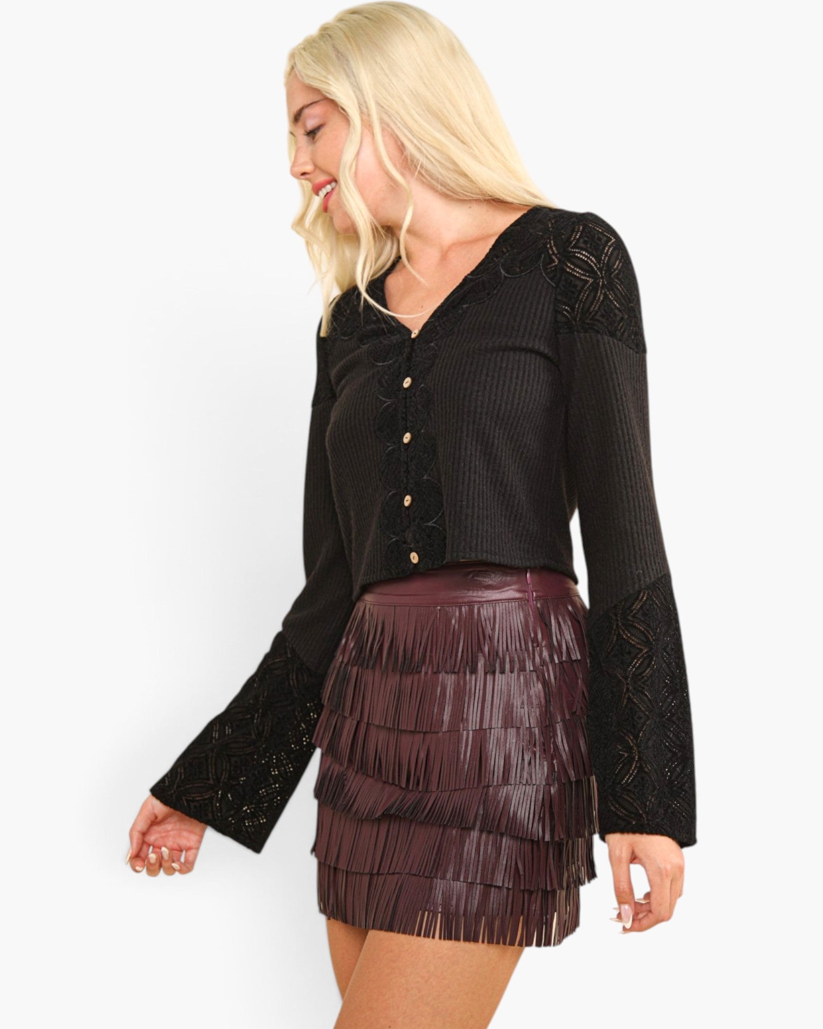 A blonde woman wearing a black long-sleeve button-front top with lace sleeves and a dark burgundy fringe mini skirt, photographed against a plain white studio background.