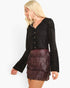 A blonde woman wearing a black long-sleeve button-front top with lace sleeves and a dark burgundy fringe mini skirt, photographed against a plain white studio background.