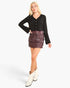 A woman modeling a black long-sleeve button-front top and a dark brown tiered fringe mini skirt, paired with white cowboy boots, against a light gray studio background.