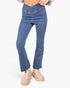 A person wearing medium-wash blue denim jeans with a flared bootcut or bootcut flare, paired with beige ankle boots and a patterned top, shown against a plain light gray studio background.