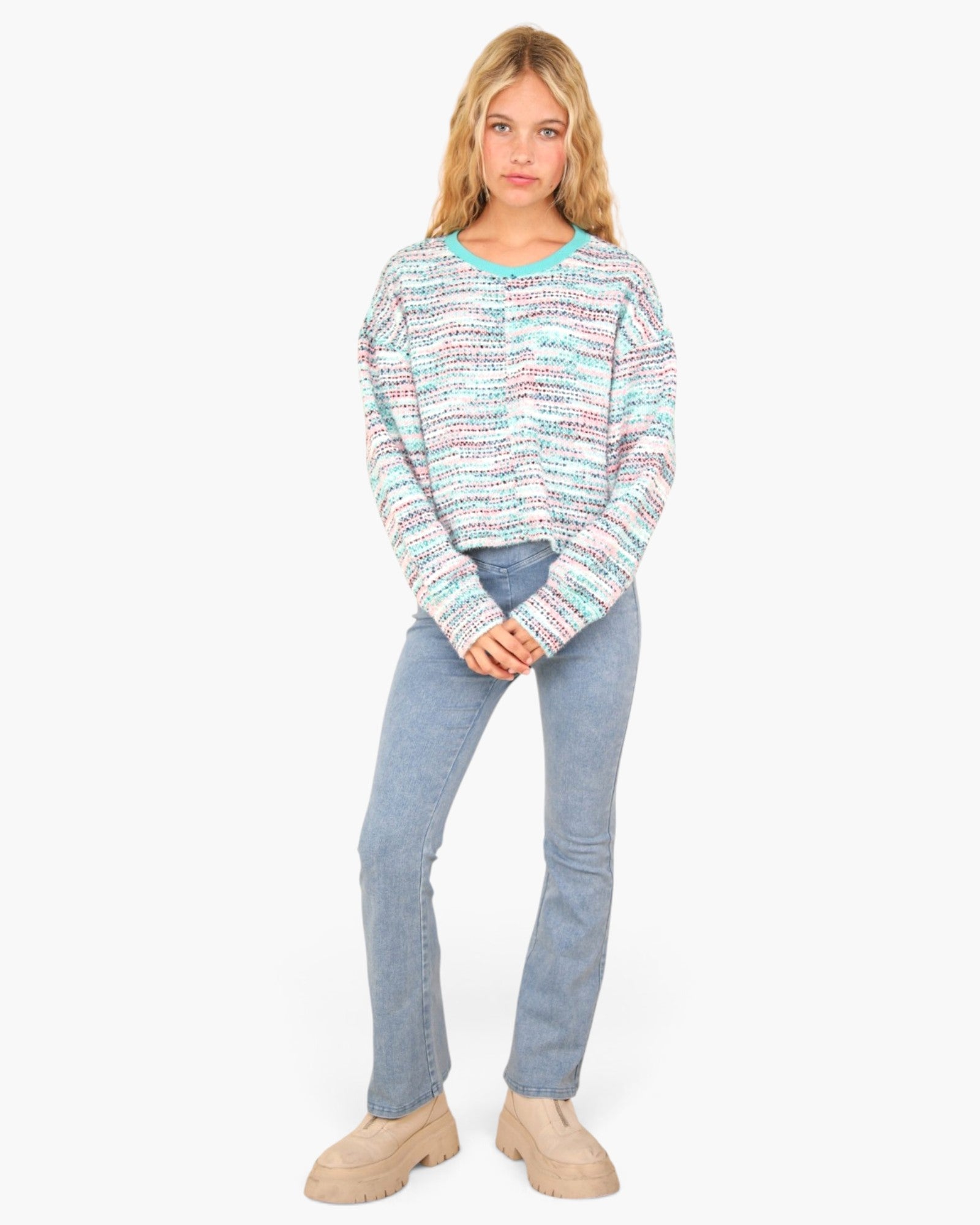 A woman modeling a long-sleeve knit sweater with a multicolor horizontal stripe pattern in shades of blue, pink, and white, paired with light-wash blue jeans and beige boots against a plain white background.