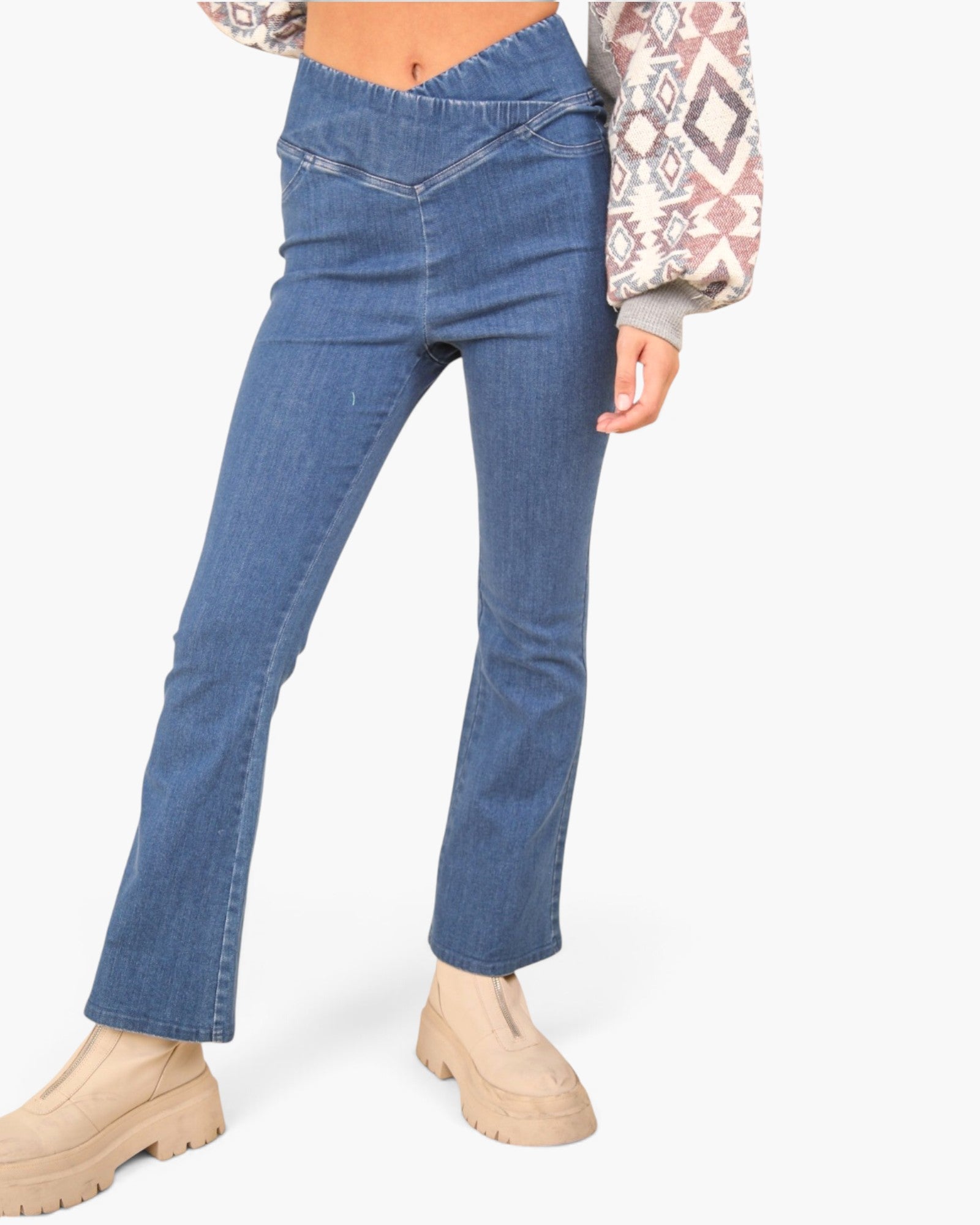 A model wearing a pair of medium-wash blue denim flare jeans with a raw, frayed hem, styled with beige platform boots and a patterned long-sleeve top against a light gray background.