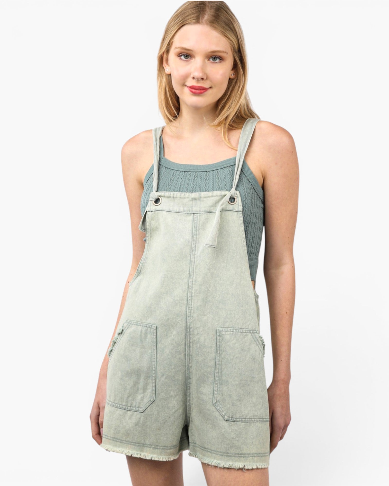 A woman modeling a light sage green denim overall romper with frayed hems, front pockets, and adjustable shoulder straps, worn over a muted blue-green ribbed tank top against a plain white background.