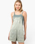A woman modeling a light sage green denim overall romper with frayed hems, front pockets, and adjustable shoulder straps, worn over a muted blue-green ribbed tank top against a plain white background.